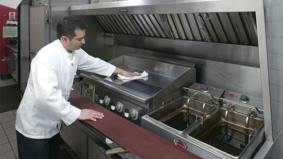 Chef cleaning a stainless-steel grill from grease residue in a commercial kitchen.