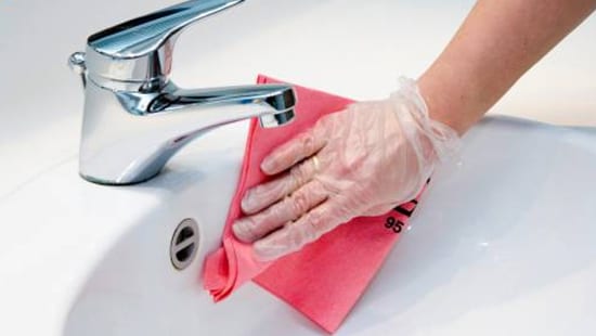 hand wearing plastic gloves sanitizing and disinfecting a porcelain sink with a cleaning rag