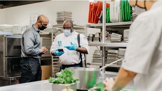 African American Service Rep meeting with restaurant manager in kitchen using Ecolab Sales & Service on phone.