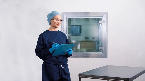 Person in cleanroom attire writes on a clipboard beside a sterile lab window and metal table.